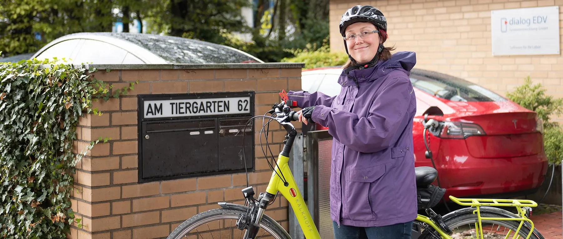 Mitarbeiterin mit Fahrrad vor dem Firmenschild Am Tiergarten 62, daneben ein Elektroauto an der Ladesäule